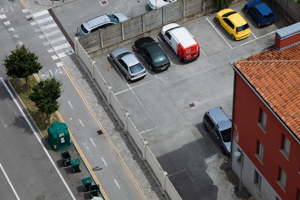 An aerial view of a parking lot with several cars parked in designated spots. The lot is adjacent to a building with a terracotta roof. A street runs alongside the parking lot, featuring bicycle lanes and waste bins lined up near the sidewalk. Trees border the road, providing green accents to the scene.