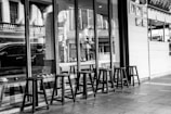 A series of wooden stools arranged in a row outside a storefront with large glass windows. The setting appears to be urban, with reflections of buildings and a car visible in the glass. The interior of the shop includes a visible counter and hanging decorations.