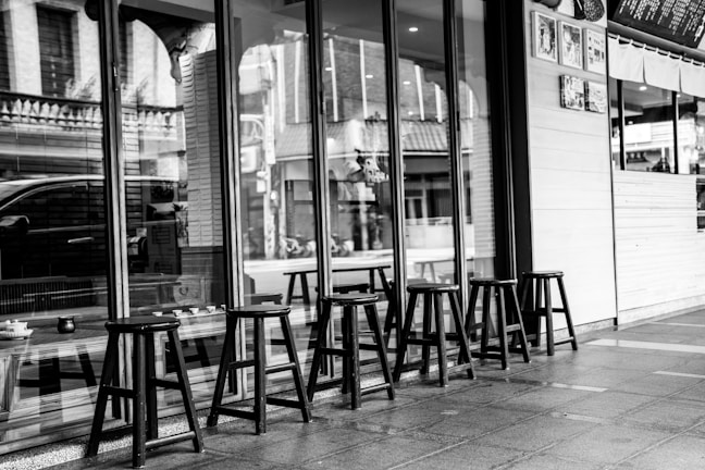 A series of wooden stools arranged in a row outside a storefront with large glass windows. The setting appears to be urban, with reflections of buildings and a car visible in the glass. The interior of the shop includes a visible counter and hanging decorations.