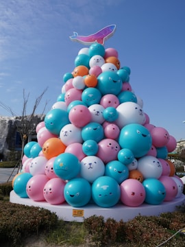 A large sculpture composed of colored balloons arranged in a conical shape, resembling a playful Christmas tree. The balloons feature cute animal faces in a variety of expressions, with pastel shades of teal, pink, white, and orange. A stylized pink and blue whale is placed on top of the structure, contrasting against a clear blue sky. The base is surrounded by a neatly trimmed hedge, adding greenery to the scene.