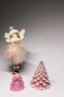 A decorative angel figurine with fluffy white wings and a soft, feathered pink dress stands alongside a small pink candle shaped like a stylized Christmas tree. A smaller pink soap or candle shaped like an angel is located in the foreground.