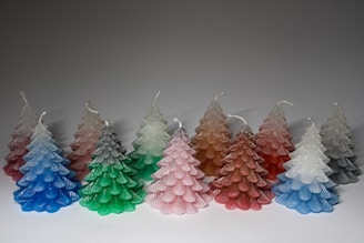 A group of candles shaped like Christmas trees are arranged in a row. They come in various colors, including pink, green, blue, red, and white. Each candle features a textured surface with layered, scalloped details resembling tree branches.