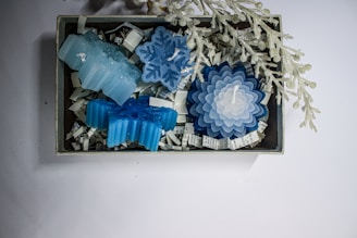 A collection of blue and white candles in various shapes, including snowflakes and flowers, displayed in a small box filled with shredded paper. Above the box, there is a decorative branch with frosted berries.