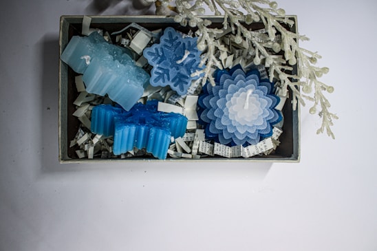 A collection of blue and white candles in various shapes, including snowflakes and flowers, displayed in a small box filled with shredded paper. Above the box, there is a decorative branch with frosted berries.