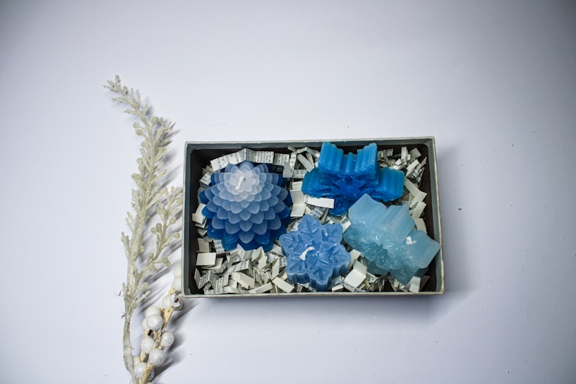 A box contains four blue decorative candles in various shapes, such as a flower, a snowflake, and abstract designs. The candles rest on shredded paper that includes black and white print. To the left of the box, there is a decorative branch with frosted white berries and leaves.
