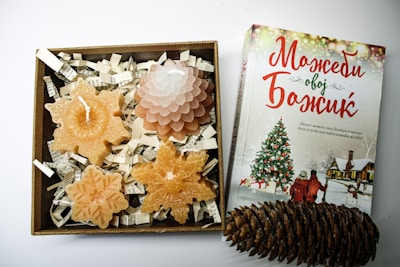 A neatly arranged gift box containing decorative candles shaped like snowflakes and a pine cone sits next to a book with a holiday theme cover featuring a Christmas tree and figures in winter attire. The box includes shredded paper as filler.