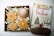 A neatly arranged gift box containing decorative candles shaped like snowflakes and a pine cone sits next to a book with a holiday theme cover featuring a Christmas tree and figures in winter attire. The box includes shredded paper as filler.