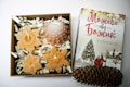 A neatly arranged gift box containing decorative candles shaped like snowflakes and a pine cone sits next to a book with a holiday theme cover featuring a Christmas tree and figures in winter attire. The box includes shredded paper as filler.