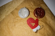 A flatlay of two round candles, one white and one dark orange, each with a face in relief on their surface. They rest on brown paper sprinkled with tiny glitter specks. Below the candles is a red and white mitten ornament with a small string attached.