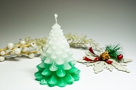 A candle shaped like a pine tree with a gradient from white at the top to green at the bottom. In the background, there are decorative elements including white berries on golden branches and a small pine cone accented with green pine needles, red berries, and a red plaid bow.