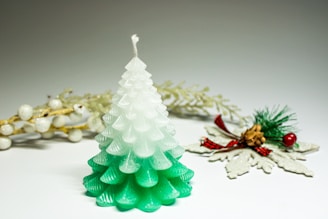 A candle shaped like a pine tree with a gradient from white at the top to green at the bottom. In the background, there are decorative elements including white berries on golden branches and a small pine cone accented with green pine needles, red berries, and a red plaid bow.