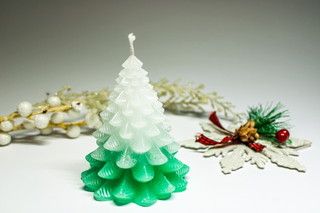 A candle shaped like a pine tree with a gradient from white at the top to green at the bottom. In the background, there are decorative elements including white berries on golden branches and a small pine cone accented with green pine needles, red berries, and a red plaid bow.