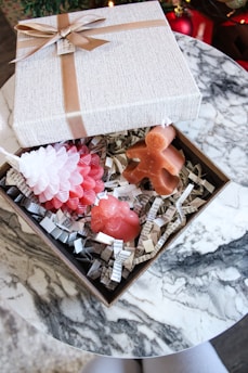 An elegant gift box with a beige texture and brown ribbon sits open on a marble table. Inside the box, there are three festive candles shaped like a pinecone, a gingerbread man, and a bear, surrounded by shredded paper strips with text.