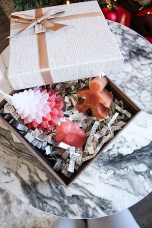 An elegant gift box with a beige texture and brown ribbon sits open on a marble table. Inside the box, there are three festive candles shaped like a pinecone, a gingerbread man, and a bear, surrounded by shredded paper strips with text.