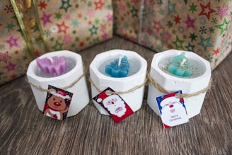 Three white candle holders with holiday-themed candles are arranged on a wooden surface. Each holder is adorned with a small card attached by twine, featuring festive designs including a reindeer, Santa Claus, and Christmas greetings. The background includes wrapped gifts with colorful star patterns.