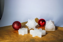 A set of decorative white candles shaped like stars and other festive forms are arranged on a brown surface. Two glossy red ornaments accompany the candles, adding a vibrant contrast to the scene.
