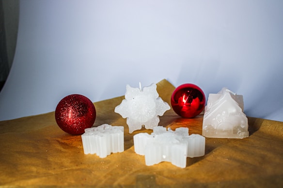 A set of decorative white candles shaped like stars and other festive forms are arranged on a brown surface. Two glossy red ornaments accompany the candles, adding a vibrant contrast to the scene.