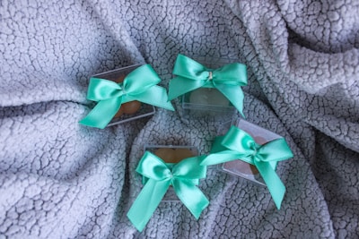 Four small transparent boxes are placed on a textured gray fabric. Each box is adorned with a mint green satin bow tied on top. The soft and cozy fabric provides a contrasting backdrop to the glossy bows and boxes.