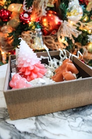 A gift box contains festive-shaped candles, including a pink and white tree and other red and brown shapes, surrounded by shredded paper filling. The box sits on a marble surface, with a decorated Christmas tree adorned with red and gold ornaments and string lights in the background.