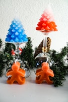 Two tree-shaped candles, one blue and one red, are placed on inverted wine glasses with green foliage arranged around them. In front of each glass is an orange gingerbread man-shaped candle, set against a plain white background and a pine cone accent.