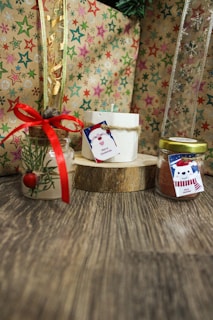 A wooden surface holds a collection of decorative items, including a candle with a 'Merry Christmas' tag tied with twine, placed on a wooden coaster. To its left, there's a small jar adorned with pine sprigs and a red ribbon. On the right, another jar with a yellow cap features a 'Merry Christmas' tag with a festive snowman illustration. The background consists of wrapping paper with colorful stars and snowflakes, creating a holiday atmosphere.