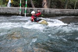 A person wearing a helmet and red jacket is paddling a kayak through white water rapids. The scene is set in an outdoor environment with a concrete barrier and trees in the background. The water is turbulent, creating an adventurous and dynamic action scene.