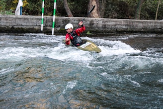 A person wearing a helmet and red jacket is paddling a kayak through white water rapids. The scene is set in an outdoor environment with a concrete barrier and trees in the background. The water is turbulent, creating an adventurous and dynamic action scene.