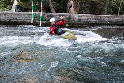 A person wearing a helmet and red jacket is paddling a kayak through white water rapids. The scene is set in an outdoor environment with a concrete barrier and trees in the background. The water is turbulent, creating an adventurous and dynamic action scene.