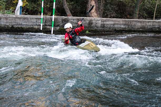 A person wearing a helmet and red jacket is paddling a kayak through white water rapids. The scene is set in an outdoor environment with a concrete barrier and trees in the background. The water is turbulent, creating an adventurous and dynamic action scene.