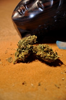 There is a wooden mask with a serene face laying on its side on a sandy surface. In front of the mask, there are a few pieces of dried cannabis buds. The setting has a warm, earthy tone with a focus on the natural elements.