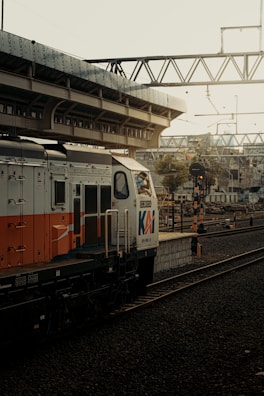 A train is positioned on railway tracks at a station. There is an overhead pedestrian bridge and numerous wires above. The train prominently displays branding, and there are signal lights along the tracks. The environment appears industrial and somewhat aged, with muted colors indicating an early morning or late afternoon setting.