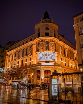 A grand theater building adorned with intricate architectural details and warm lighting, prominently displaying a sign for 'The Book of Mormon'. The scene is set at night with the wet pavement reflecting the city lights, and a bus and people are visible in the foreground under umbrellas.