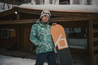 A person in winter sports gear stands outside a wooden cabin, holding a snowboard. The outfit is a green and blue pixelated jacket with dark pants, and a white helmet with goggles is worn. Snow covers the ground and the cabin roof.
