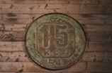 A vintage coin displaying the denomination of 15 sits atop a rustic wooden surface. The coin, marked with weathered textures, bears the year 1943 and shows signs of age with a greenish, oxidized patina. The wooden backdrop features darkened knots and varying shades of brown, contributing to an antique, historical feel.