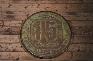 A vintage coin displaying the denomination of 15 sits atop a rustic wooden surface. The coin, marked with weathered textures, bears the year 1943 and shows signs of age with a greenish, oxidized patina. The wooden backdrop features darkened knots and varying shades of brown, contributing to an antique, historical feel.