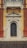 A detailed, ornate architectural entrance featuring elaborate carvings surrounding an arched wooden door. Above the door, an inscription reads 'Donazione Senatore Pietro Canonica.' The structure combines various elements including a sturdy base, intricate designs, and a small balcony with decorative latticework above, all set against a contrasting yellow wall with red brick accents.