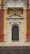 A detailed, ornate architectural entrance featuring elaborate carvings surrounding an arched wooden door. Above the door, an inscription reads 'Donazione Senatore Pietro Canonica.' The structure combines various elements including a sturdy base, intricate designs, and a small balcony with decorative latticework above, all set against a contrasting yellow wall with red brick accents.