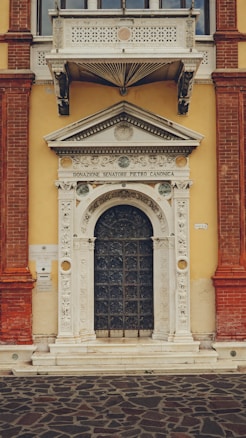 A detailed, ornate architectural entrance featuring elaborate carvings surrounding an arched wooden door. Above the door, an inscription reads 'Donazione Senatore Pietro Canonica.' The structure combines various elements including a sturdy base, intricate designs, and a small balcony with decorative latticework above, all set against a contrasting yellow wall with red brick accents.