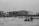 A black and white image of a beach with several people engaging in leisure activities. In the background, there is a large unfinished building with cranes, and some trees line the shore. Distant city buildings are visible on the horizon.