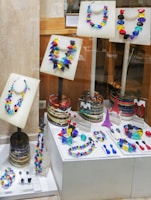 A collection of vibrant, multicolored necklaces and earrings displayed on mannequins within a store window. The pieces of jewelry are arranged on white stands and platforms, showcasing an array of geometric shapes and bright colors. The background consists of wooden and stone surfaces, adding a contrasting texture to the display.