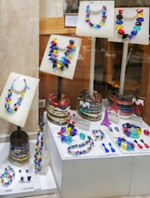 A collection of vibrant, multicolored necklaces and earrings displayed on mannequins within a store window. The pieces of jewelry are arranged on white stands and platforms, showcasing an array of geometric shapes and bright colors. The background consists of wooden and stone surfaces, adding a contrasting texture to the display.
