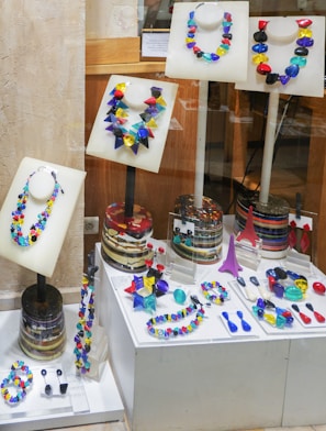 A collection of vibrant, multicolored necklaces and earrings displayed on mannequins within a store window. The pieces of jewelry are arranged on white stands and platforms, showcasing an array of geometric shapes and bright colors. The background consists of wooden and stone surfaces, adding a contrasting texture to the display.