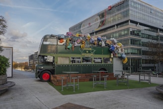 A vintage double-decker bus painted in dark green sits on an outdoor patio area. The top of the bus is covered with a variety of objects and colorful items, such as stuffed toys and other playful decorations. Surrounding the bus are outdoor tables and benches resting on a patch of artificial grass. The background features a modern glass office building under a partly cloudy sky.