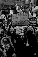 A black-and-white photograph of a street protest featuring many people holding signs and flags. The prominent sign in the center reads 'Stop Appeasing Genocide. It didn't work in 1938. It won't work now.' Many other signs read 'Freedom for Palestine' and 'End the Occupation Now.' The crowd is diverse, with people of various ages and ethnic backgrounds.