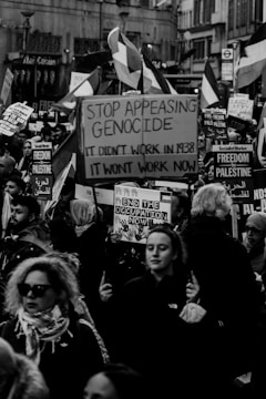 A black-and-white photograph of a street protest featuring many people holding signs and flags. The prominent sign in the center reads 'Stop Appeasing Genocide. It didn't work in 1938. It won't work now.' Many other signs read 'Freedom for Palestine' and 'End the Occupation Now.' The crowd is diverse, with people of various ages and ethnic backgrounds.