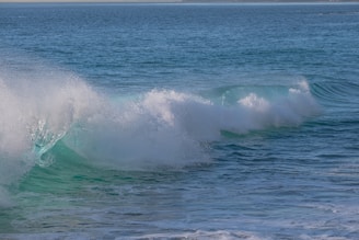 A large ocean wave is captured in mid-crash, with foamy white water at the crest and deep turquoise below. The surface of the water is textured and rippling, extending out to a distant horizon.
