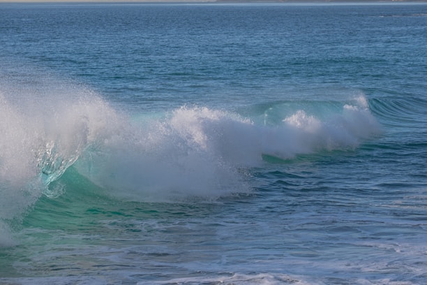 A large ocean wave is captured in mid-crash, with foamy white water at the crest and deep turquoise below. The surface of the water is textured and rippling, extending out to a distant horizon.