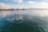 A person is paddleboarding on a large, calm body of water with gentle ripples. In the distance, there are trees and a slightly snowy landscape under a partly cloudy sky. A sailboat is also visible further away, adding to the serene and peaceful setting.
