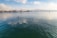 A person is paddleboarding on a large, calm body of water with gentle ripples. In the distance, there are trees and a slightly snowy landscape under a partly cloudy sky. A sailboat is also visible further away, adding to the serene and peaceful setting.