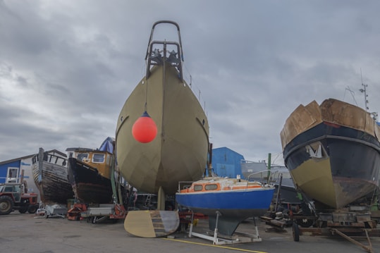 A shipyard scene with several boats on land, including a large boat with a bright red buoy hanging from it in the center. On the left, there is an older wooden boat, and on the right, a black and brown vessel. A small blue sailboat is in front, and various equipment and a red tractor can be seen in the foreground, with a cloudy sky above.